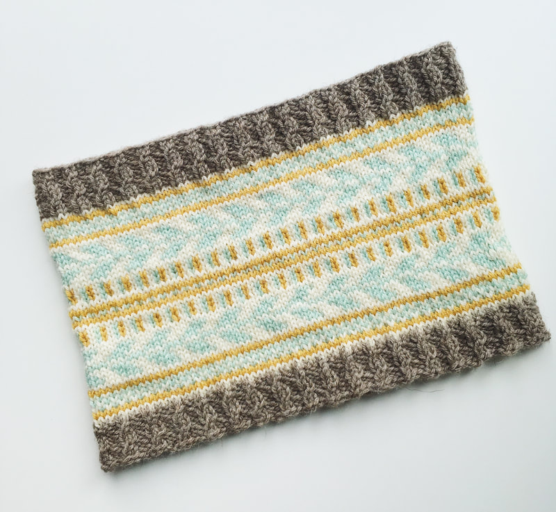 A cowl is shown laid flat on a white background. The cowl incorporates 3 colour stranded knitting and borders of brown cable ribbing. 