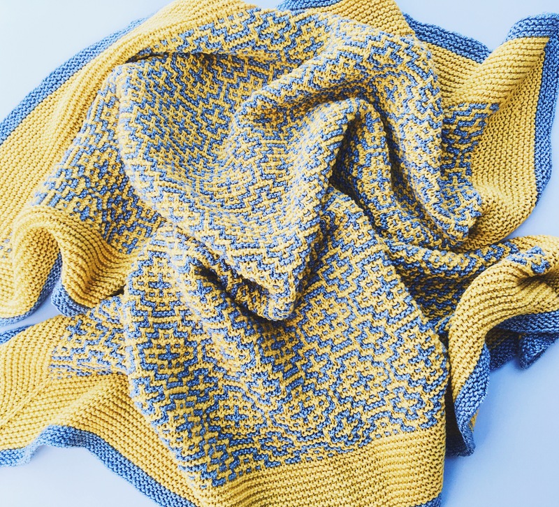 A yellow and grey blanket is pictured in a loose pile on a grey background. The blanket is textured and incorporates a tiled pattern.