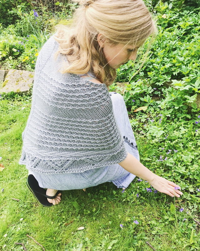 A blonde woman crouches down to pick a flower, holding her grey shawl around her shoulders. The shawl is textured and has a lace edging. 