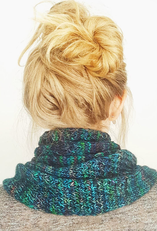 A brightly coloured cowl in shades of green and blue is wrapped three times around the neck of a blonde woman, seen from behind with her hair piled on top of her head.