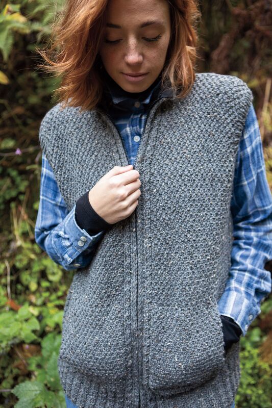 A textured vest in dark grey wool is modelled with a blue tartan shirt. The model is in the process of pulling down the zipper. 